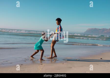 Vista laterale della giovane donna afroamericana che tiene le mani della figlia e gioca in spiaggia contro il cielo Foto Stock