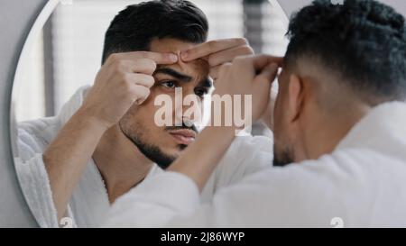 Infelice arabo indiano arabo indiano uomo bearded guardando nello specchio della stanza da bagno sensibilità preoccupata circa il problema della pelle facciale acne. Ragazzo ansioso millennial che stringe Foto Stock