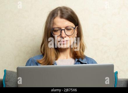Una giovane donna lavora su un computer portatile mentre si siede sul divano di casa. Foto Stock