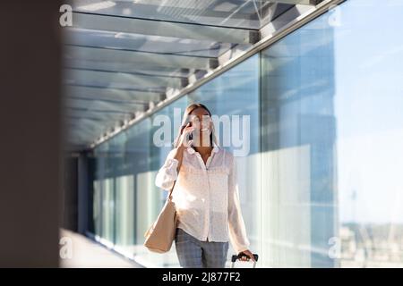 Sorridente donna d'affari biraciale che parla su smartphone camminando con i bagagli nel corridoio all'aeroporto Foto Stock