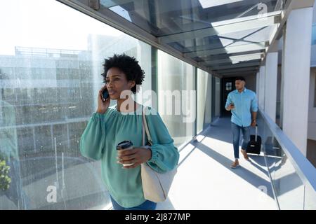 Donna d'affari afroamericana che parla sullo smartphone mentre uomo d'affari cammina con i bagagli Foto Stock