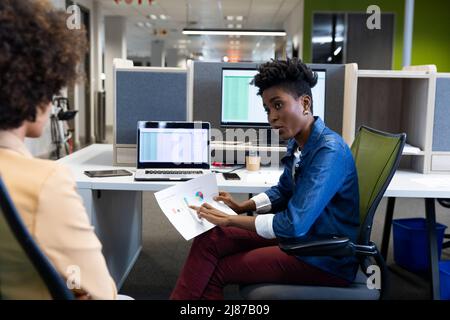 Fiduciosa donna d'affari afroamericana che spiega documento a una collega ispanica in cubicolo Foto Stock