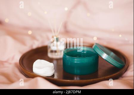 Maschera facciale in gel idratante in vaso di plastica con tamponi in cotone e profumo liquido in bottiglia di vetro su vassoio di legno in letto sopra le luci. Prodotti di bellezza a h Foto Stock