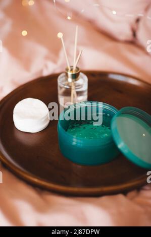 Maschera facciale in gel idratante in vaso di plastica con tamponi in cotone e profumo liquido in bottiglia di vetro su vassoio di legno in letto sopra le luci. Prodotti di bellezza a h Foto Stock