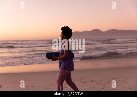 Vista laterale di donna matura afroamericana con capelli afro che trasportano stuoia yoga sulla spiaggia al tramonto Foto Stock