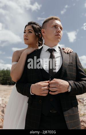 Elegante sposo in un vestito e una bella sposa in un abito bianco che si posa sullo sfondo della natura, cielo, colline e rocce. Foto di alta qualità Foto Stock