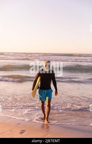 L'intera lunghezza di uomo maggiore afro-americano con la tavola da surf che cammina contro il mare e il cielo limpido Foto Stock