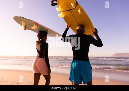 Coppia afroamericana che porta tavole da surf sulla testa che cammina verso il mare contro il cielo limpido al tramonto Foto Stock