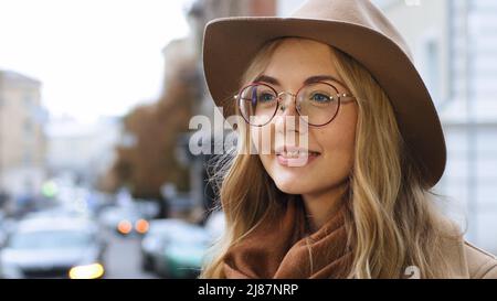 Giovane bella ragazza caucasica in piedi all'aperto close-up donna millenaria con cappello e occhiali su sfondo città ritratto sognante femmina sorridente Foto Stock