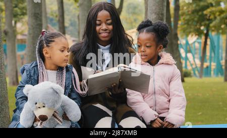 Felice famiglia afroamericana che riposa su panchina in parco giovane madre legge il libro alle figlie bambino che tiene orsacchiotto la bambina punta il dito Foto Stock