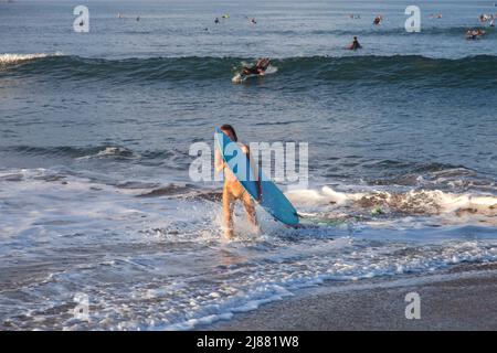 Una donna caucasica bionda che esce dall'oceano portando una lunga tavola da surf blu alla spiaggia di Batu Bolong a Canggu, Bali, Indonesia. Foto Stock