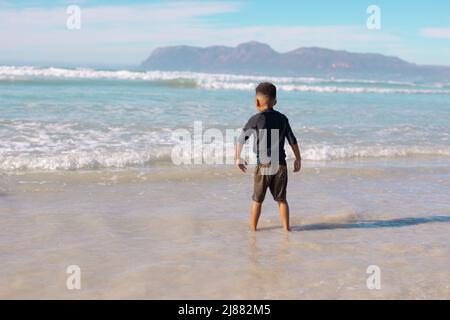 Vista posteriore del ragazzo afroamericano in piedi in mare contro il cielo durante la giornata di sole Foto Stock