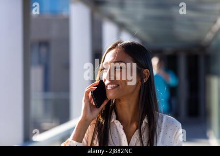 Sorridendo giovane donna d'affari biraciale che parla al telefono cellulare in corridoio sul posto di lavoro Foto Stock