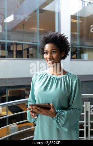 Donna d'affari afroamericana sorridente che tiene un tablet pc che guarda lontano in piedi al moderno posto di lavoro Foto Stock
