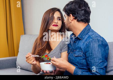 Giovane coppia indiana seduta in soggiorno e mangiare insalata di verdure Foto Stock
