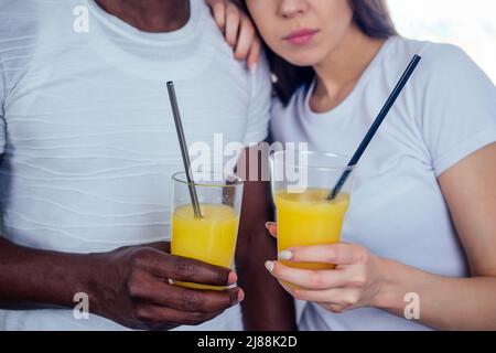 coppia internazionale in amore tenendo in mani occhiali con succo d'arancia.afro uomo utilizzare stoviglie monouso, ma donna europea using eco tubi d'acciaio Foto Stock