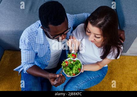 uomo afroamericano che mangia insalata con donna in soggiorno Foto Stock