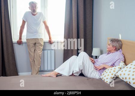 bionda shorh haircut donna litigando con il suo marito anziano in luce del giorno bedroom.handsome uomo in piedi al davanzale e guardando la moglie Foto Stock