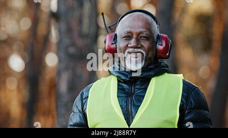 Uomo anziano ingegnere forestale professionale in cuffie protettive insonorizzate in piedi in foresta guardando fotocamera caposquadra afroamericano maturo Foto Stock
