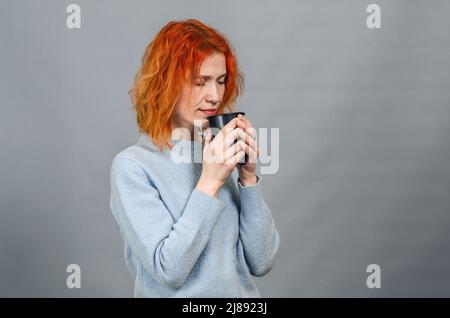 Giovane donna dai capelli rossi con bevanda a occhi chiusi dal piacere, vista laterale Foto Stock