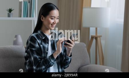Asian felice sorridente ragazza donna seduta a casa divano guardando il telefono cellulare chattare navigazione utilizzando wi-fi web service guardare video online app SMS Foto Stock