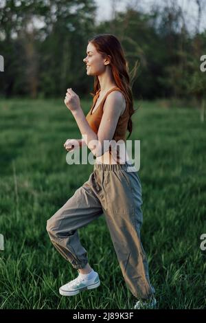 Una donna atletica scivola sull'erba in pantaloni verdi e sneakers e allena il suo corpo per la stagione estiva nel parco alla luce del sole del tramonto Foto Stock