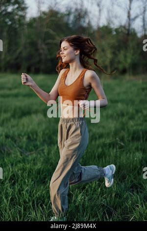 Una donna atletica scivola sull'erba in pantaloni verdi e sneakers e allena il suo corpo per la stagione estiva nel parco alla luce del sole del tramonto Foto Stock