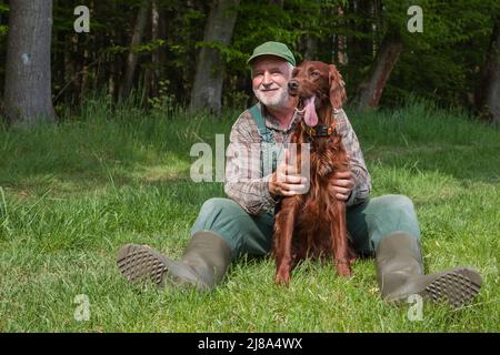 Un vecchio cacciatore in dungaree e stivali di gomma verde è seduto al sole al bordo della foresta e tiene amorevolmente il suo Irish Setter Hound . Foto Stock