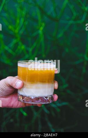 Tenere a mano il caffè latte ghiacciato in un bicchiere alto. Bevanda estiva fredda con sfondo verde naturale Foto Stock