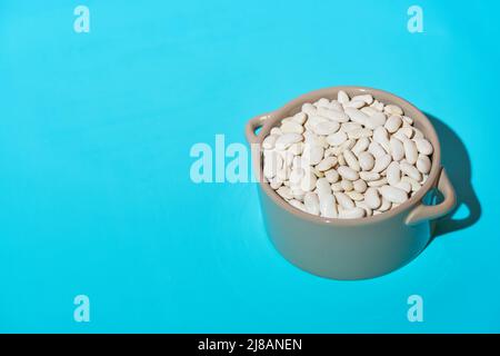Fagioli bianchi secchi in un recipiente di vetro grigio su sfondo blu chiaro. Luce dura, ombra profonda. Primo piano, spazio di copia. Foto Stock
