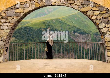 Turista femminile in Abaya e Hijāb controlla il telefono in piedi in Arch of Georgia Friendship Monument sulla Military Highway con bella vista montagna dietro Foto Stock