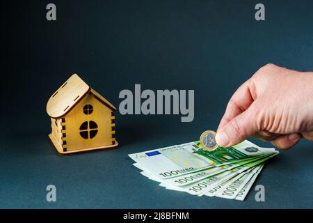 Uomo che mette una moneta di euro su cento banconote di euro di fronte a casa giocattolo in legno su sfondo grigio. Foto Stock
