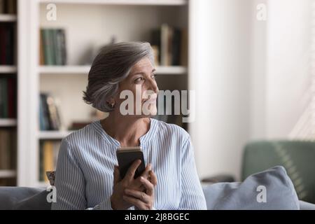 Donna utente senior positivo e pensivo seduto sul divano di casa Foto Stock