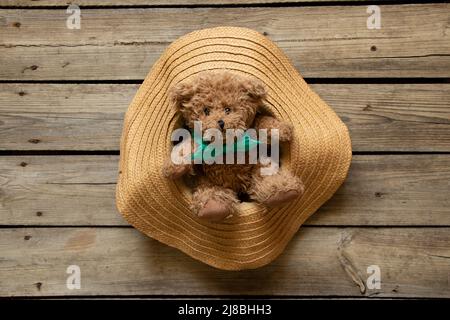 Un orsacchiotto marrone si siede in un cappello di donna paglia su un tavolo di legno, moda Foto Stock