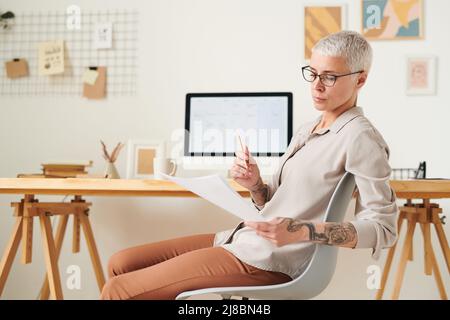 Donna centrata di mezza età in occhiali seduti in ufficio moderno e contratto di lettura Foto Stock