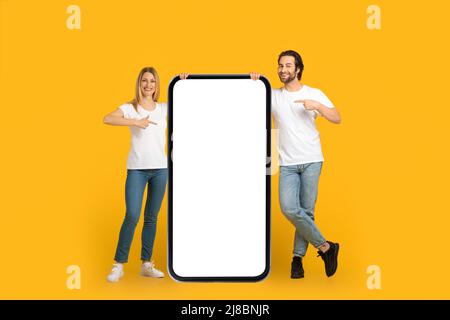 Soddisfatto coppia millennial europea in magliette bianche puntano le dita su un telefono enorme con schermo vuoto Foto Stock