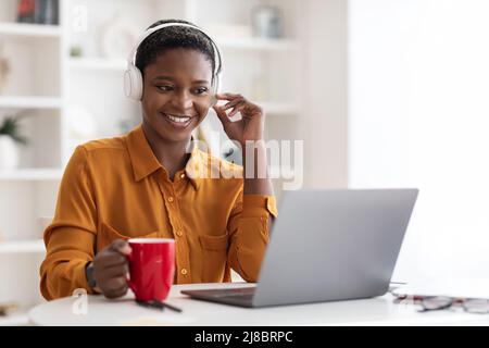 Giovane donna nera che ha una videoconferenza con i colleghi, bevendo caffè Foto Stock
