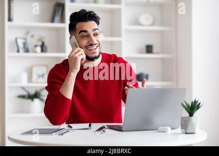 Felice giovane arabo maschio con barba parla al telefono con il cliente sul posto di lavoro con laptop in casa ufficio interno Foto Stock