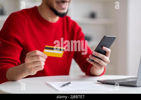 Sorridente ragazzo arabo giovane con barba che mostra la carta di credito e digita sullo smartphone in casa ufficio Foto Stock