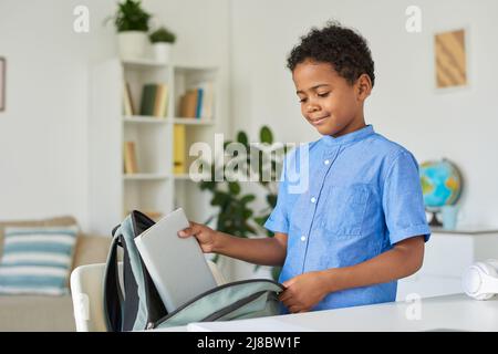 Carino ragazzo afroamericano in camicia blu che confezionano il libro in borsa mentre preparano il roba per la scuola Foto Stock