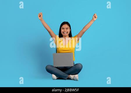 Felice giovane donna cinese in casual si siede sul pavimento con il computer portatile e solleva le mani in su gioisce nella vittoria Foto Stock