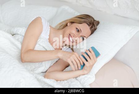 Felice bella giovane bionda europea donna legge messaggio al telefono, sdraiato sul letto in camera bianca Foto Stock