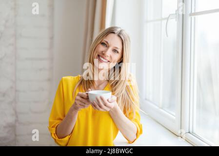 Allegra ragazza bionda europea piuttosto giovane in abiti gialli gode di tè caldo e tempo libero vicino alla finestra Foto Stock