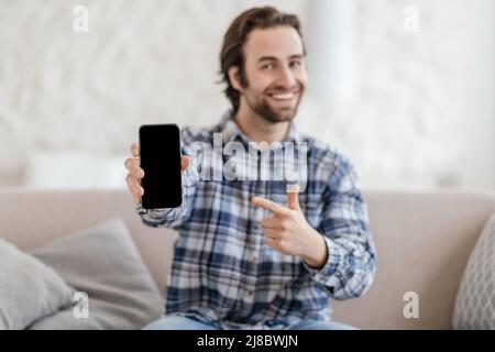 Soddisfatto maschio caucasico millenario con stoppia sedersi sul divano, puntando il dito al telefono con schermo vuoto in salotto Foto Stock