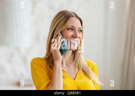 Allegro attraente ragazza bionda europea millennial in abiti gialli parla per telefono in camera interna Foto Stock