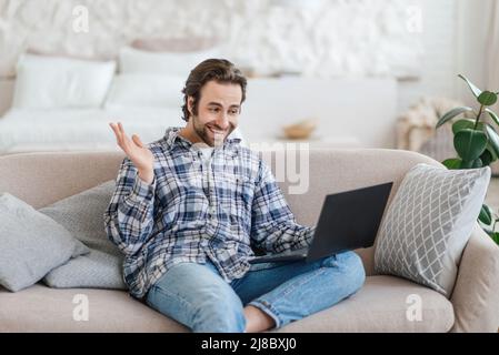 Uomo caucasico soddisfatto e millenario con stoppia si siede sul divano ondendo la mano e guardando il laptop Foto Stock