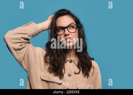 La giovane donna bruna europea pensiva in casual e occhiali graffia la testa e guarda lo spazio libero Foto Stock