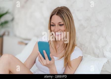 Allegro bella giovane bionda europea donna legge messaggio sul telefono in letto in bianco camera da letto interno Foto Stock