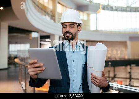 Sorridente giovane ingegnere arabo con barba in elmetto controlla il progetto su tablet e tiene i disegni Foto Stock