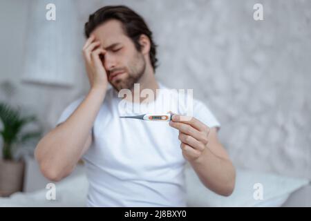 Triste disperato maschio caucasico millenario con stoppia, siede sul letto preme la sua mano alla testa e tiene termometro Foto Stock